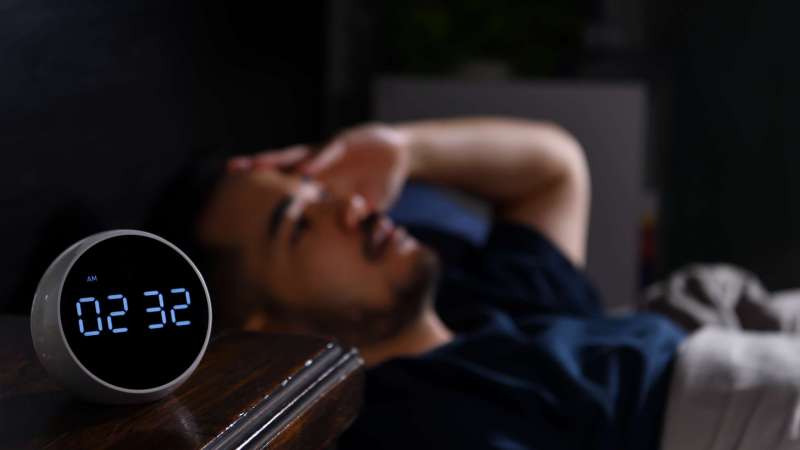 Depressed young Asian man lying in bed cannot sleep from insomnia. focus on clock